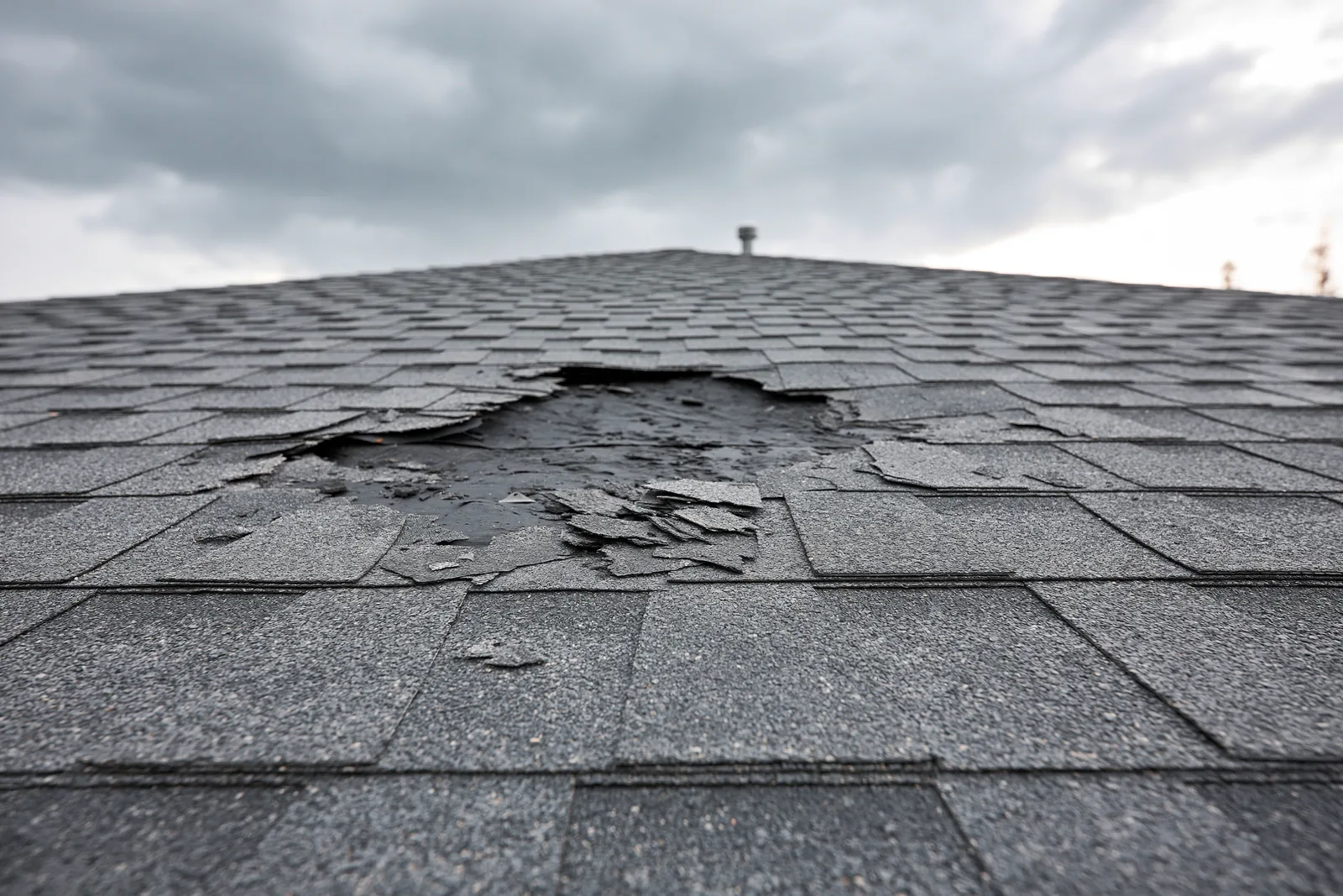 Torn shingles reveal a hole in the roof beneath a stormy sky